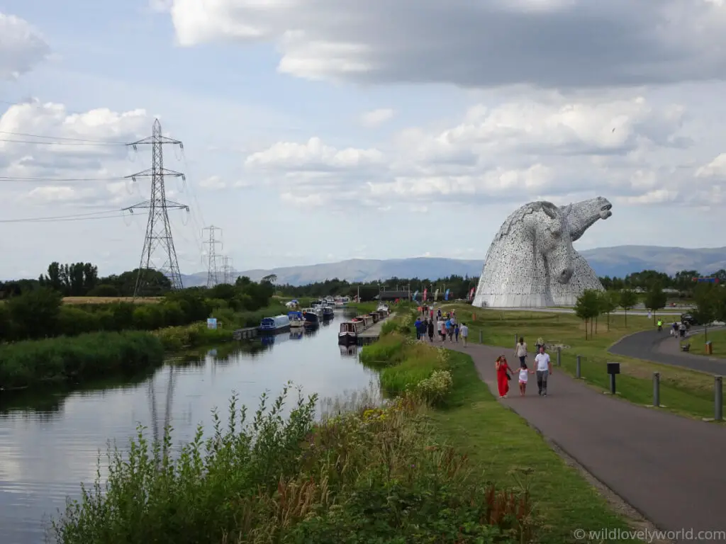 view of the forth and clyde canal with canal boats, people walking on the path alongside the canal, and the large kelpies horse head sculptures in the background