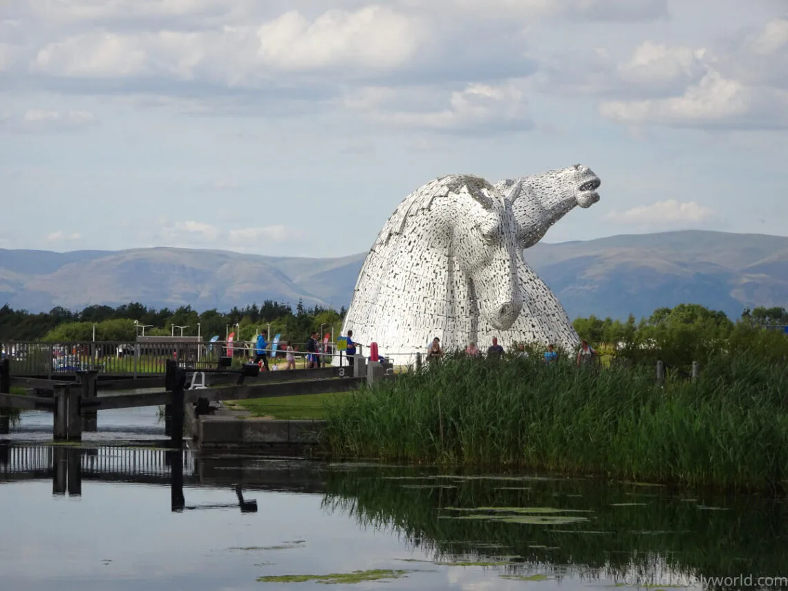 view of the kelpies from a distance, with the hills in the background, people crossing over the canal on the lock bridge walking towards the kelpies