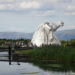 view of the kelpies from a distance, with the hills in the background, people crossing over the canal on the lock bridge walking towards the kelpies
