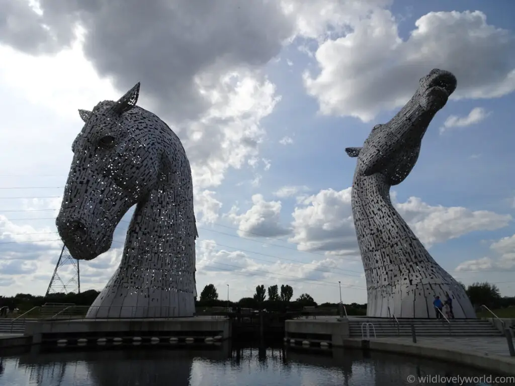 view of the kelpies from the front with the canal basin beneath and a blue sky with clouds