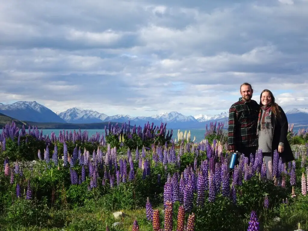 lauren and fiachra standing in a field of lupins at lake tekapo with snowy mountains in the background