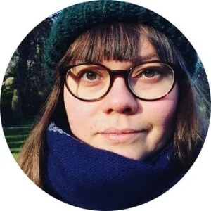 circle profile picture of lauren. she has brown hair and brown eyes, a fringe and long hair, she is wearing round brown glasses, a green hat and a blue scarf.