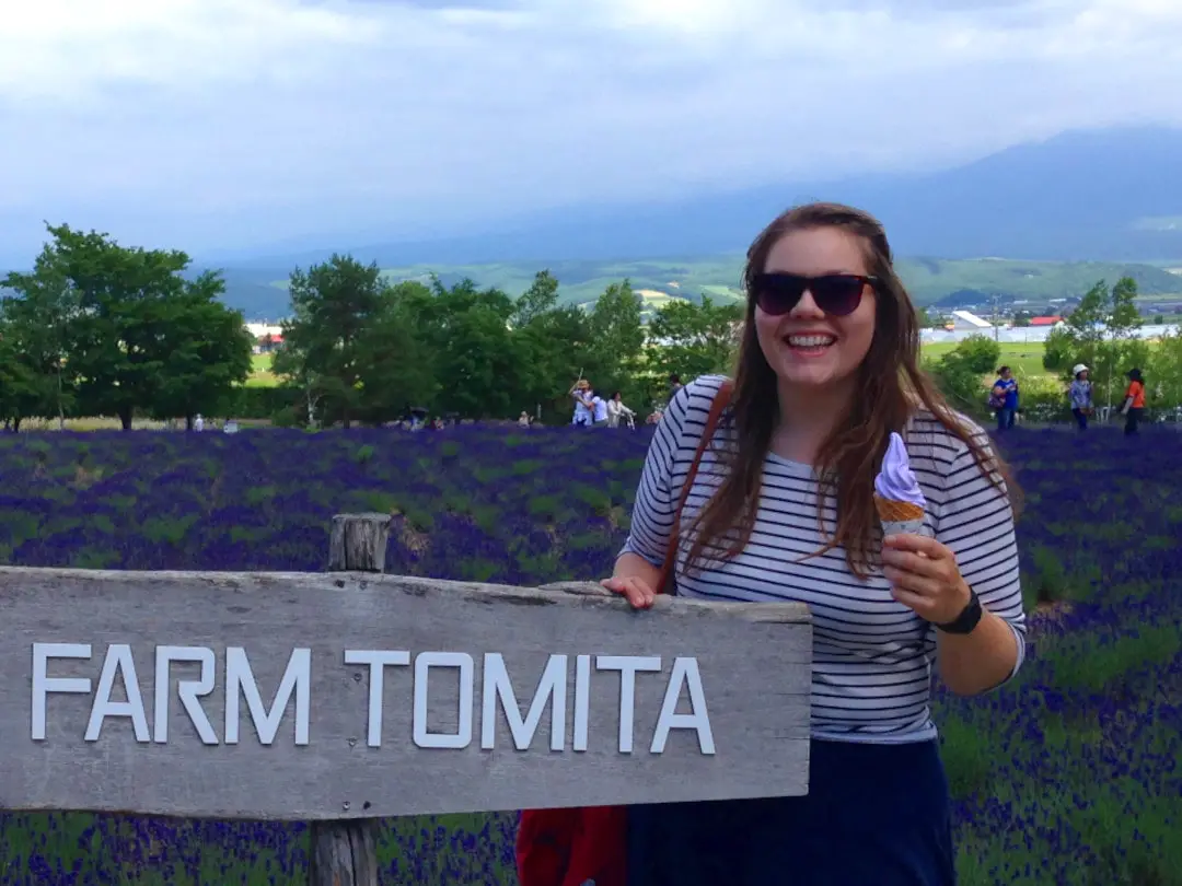 lauren standing with an ice cream in front of a lavender field next to the farm tomita sign in hokkaido