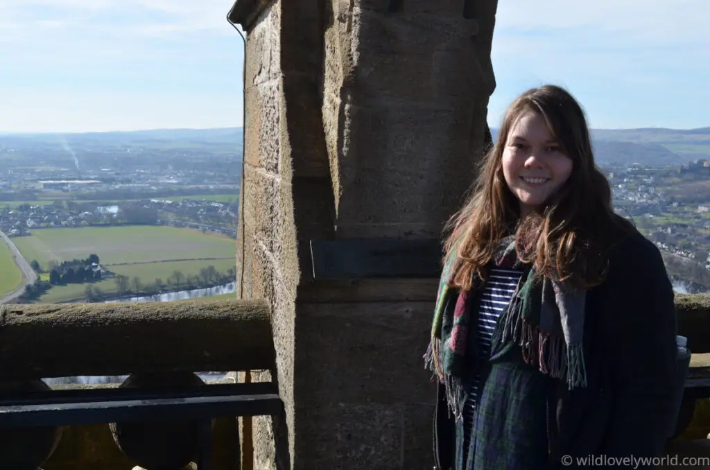 lauren smiling at the camera on top of the wallace national monument near stirling in scotland with a stone column and view of stirling and countryside in the distance