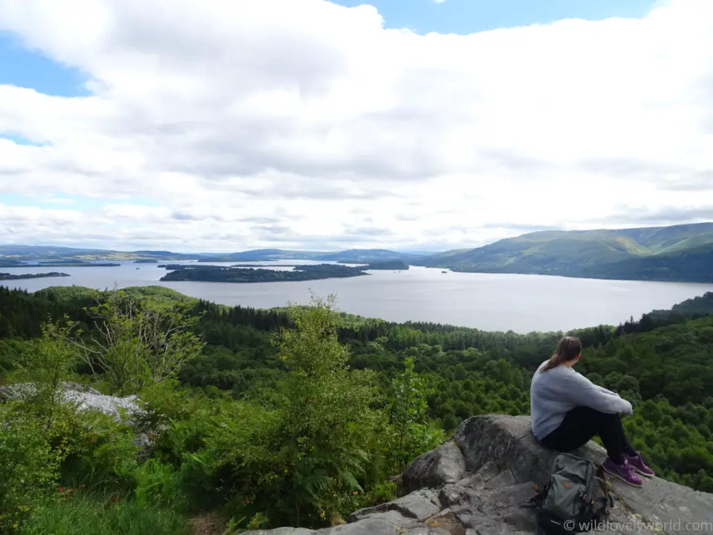 lauren sitting on a rock on a hilltop looking out at the view across loch lomond, hills and islands, in trossachs national park