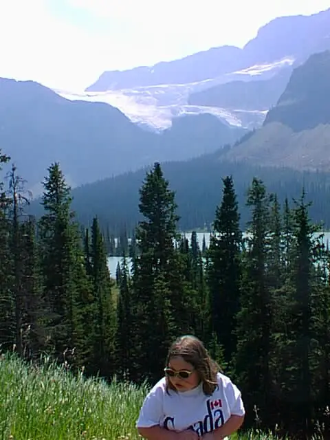 lauren as a child in alberta in canada in the 1990s. she is wearing a white t-shirt that says 'canada' with a canadian flag. behind her are green trees, a lake, and in the distance are mountains with snow and a glacier.