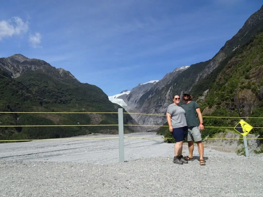 lauren and fiachra standing smiling at the camera at the fence at the viewpoint of the franz josef glacier, visible in the distance