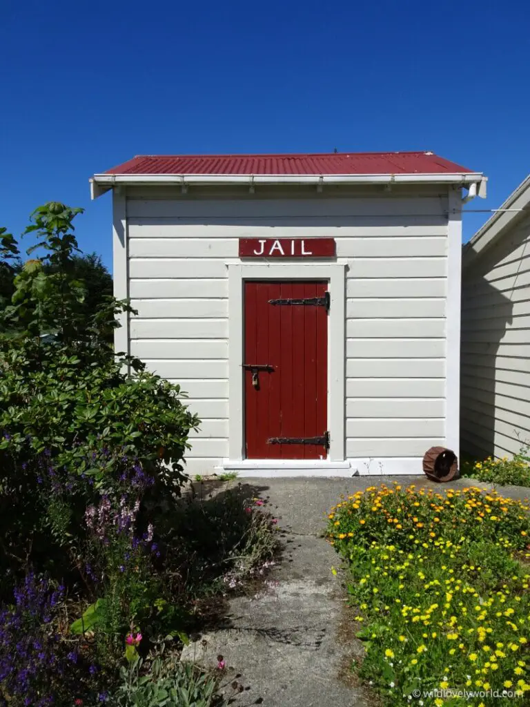 waikawa museum colonial jail house - red and white building, with a flowered garden in front and a blue sky behind