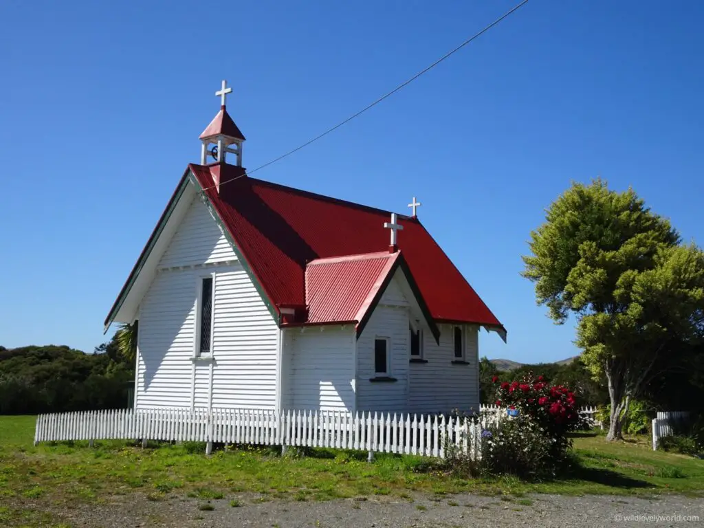 red and white wooden church with flowers, white fence, green tree, blue sky - st marys church in waikawa, new zealand