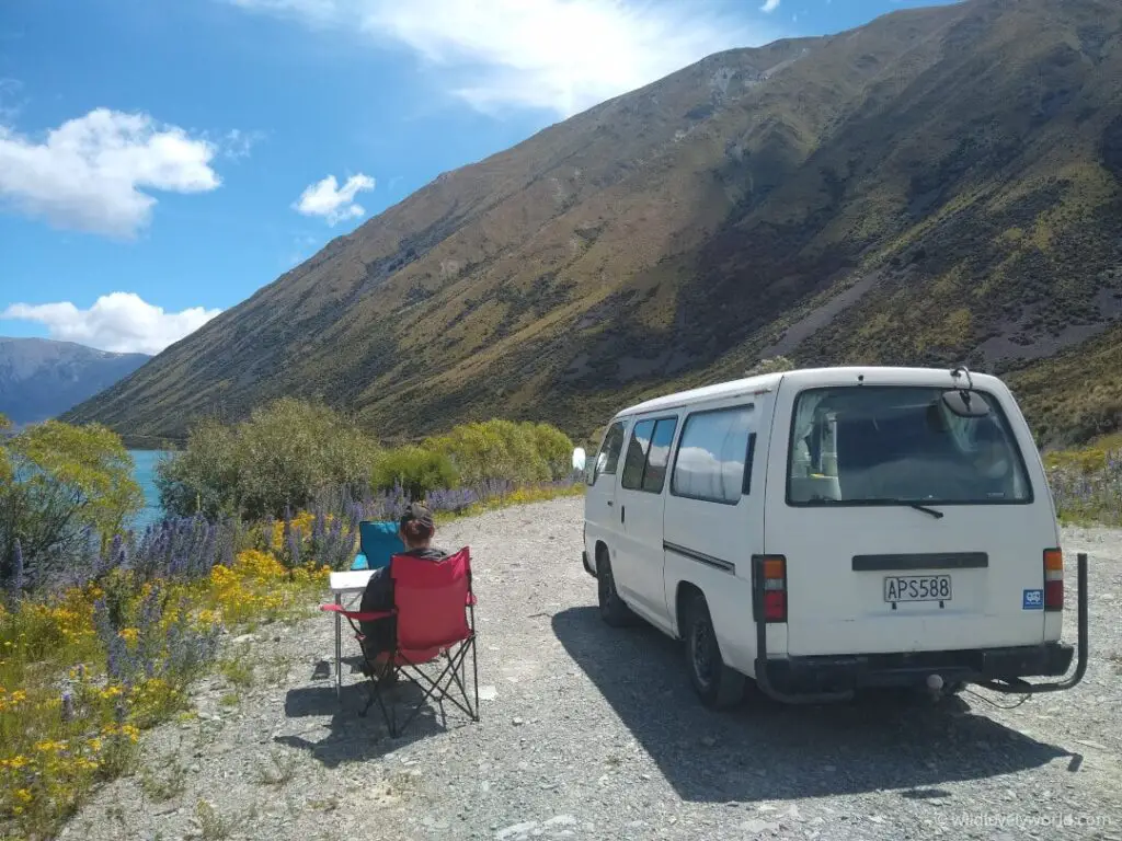 fiachra relaxing in a camping chair next to our nissan caravan campervan next to lake ohau with wild flowers on a sunny day