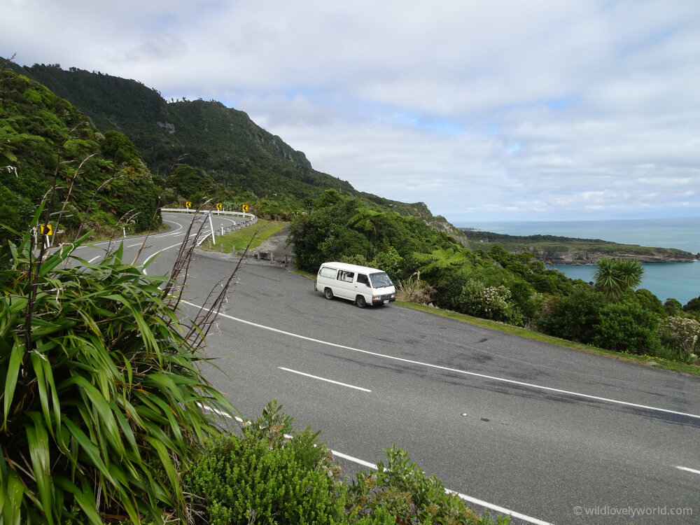 nissan caravan campervan on the side of a windy road, with lush green rainforest trees and the blue sea in the background