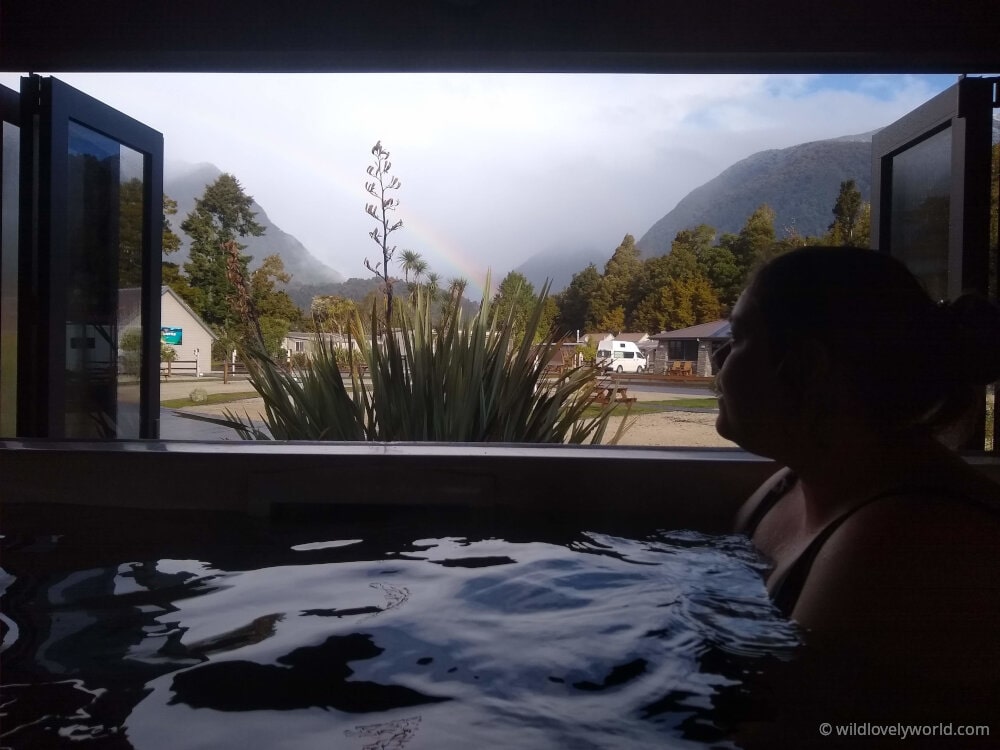 lauren enjoying the hot spa pool at fox glacier top 10 holiday park, looking out of the window at the cloudy, misty mountains and a rainbow