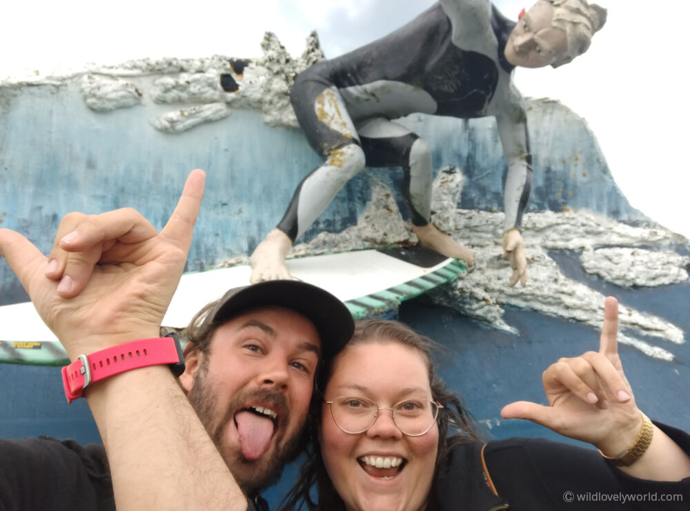 lauren and fiachra smiling and making shaka hand sign in front of the colac bay giant surfer sculpture in new zealand