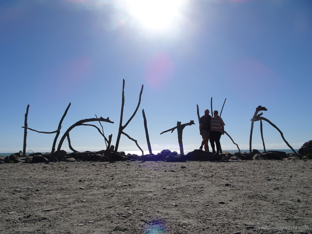 lauren and fiachra standing in front of the hokitika beach sign made of driftwood, with a blue sky and the sun high behind them