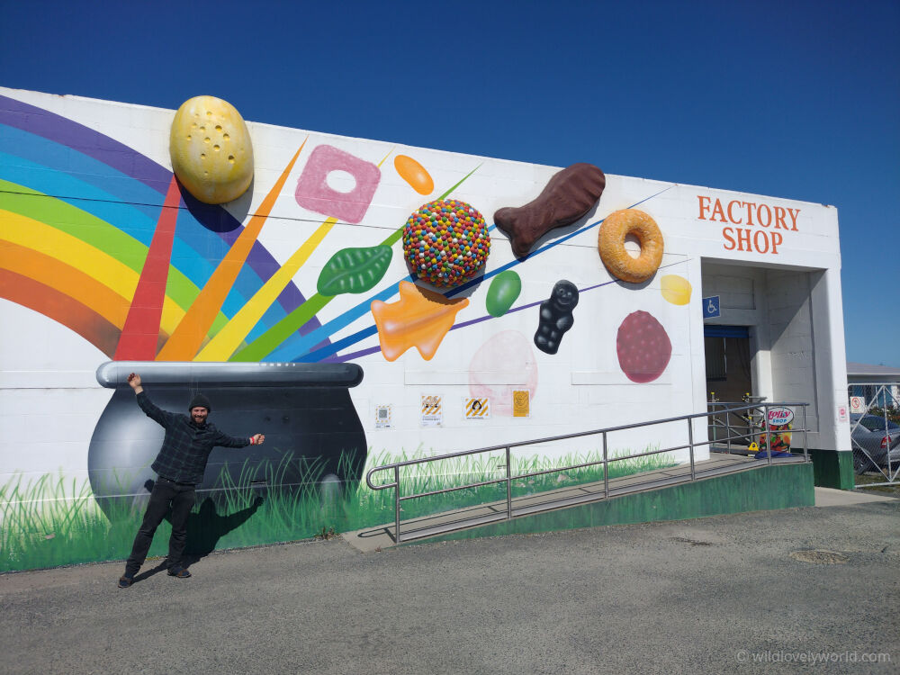 fiachra looking happy with his arms stretched out outside the rainbow confectionery factory sweet shop in oamaru new zealand. on the wall is a rainbow and giant 3D sweets