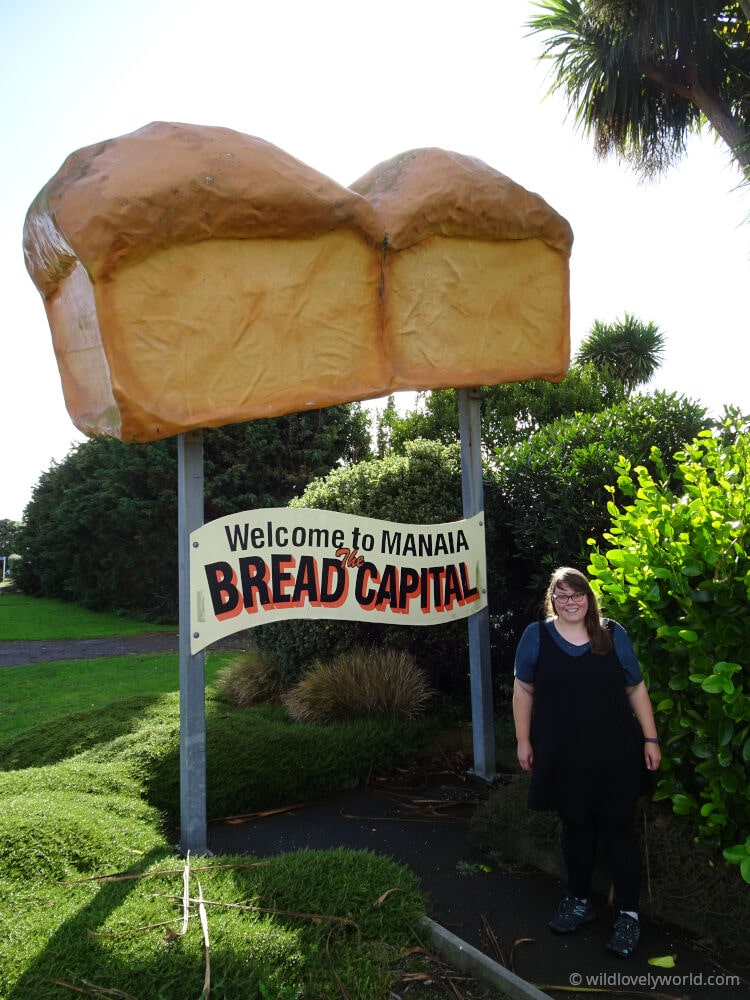 lauren standing with the welcome to manaia the bread capital sign topped with a giant loaf of bread - big things road trip new zealand