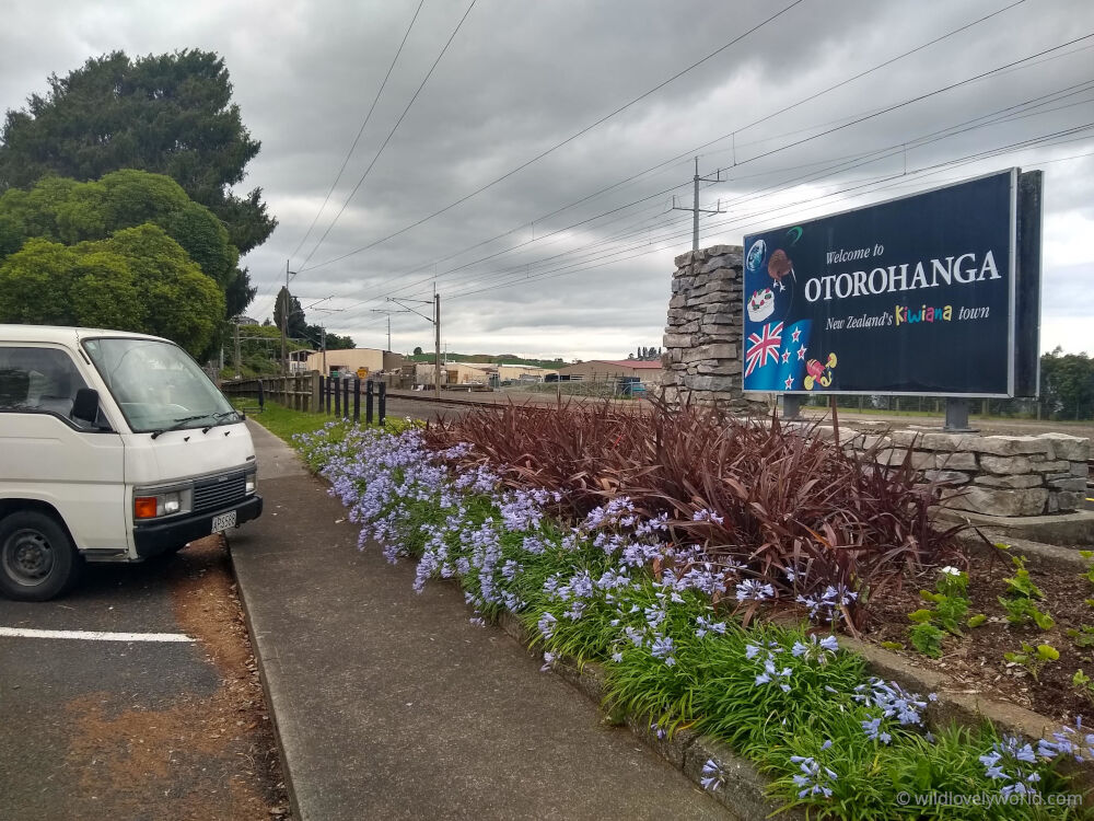 nissan caravan campervan parked next to the otorohanga railway station with flowers and a sign that says welcome to otorohanga new zealand's kiwiana town