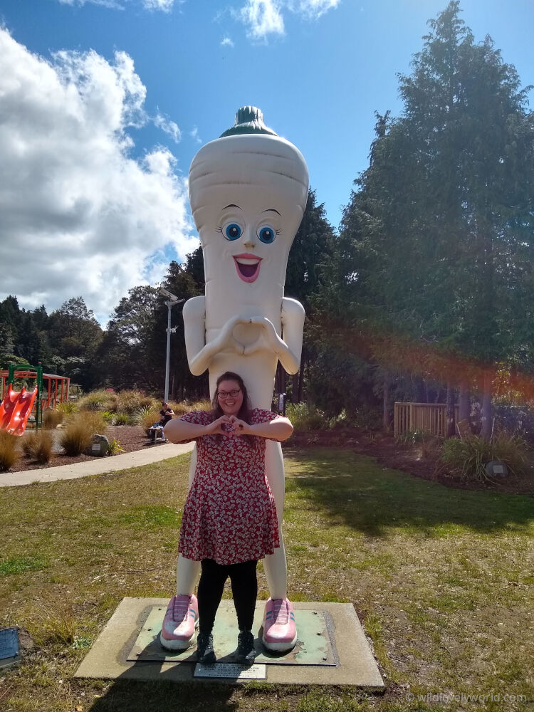 lauren standing in front of the big antropomorphic parsnip at ohakune carrot adventure park - the parsnip has big eyes and is smiling and making a heart shape with its hands - lauren is making the same heart shape