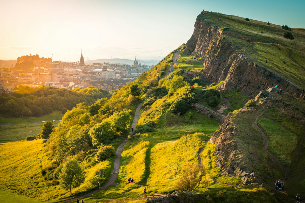 salisbury crags on the right and view of edinburgh castle and walter scott monument on the left, with golden sunlight glow coming from the left