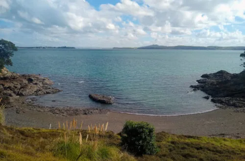 small cove beach with clear blue water, blue cloudy sky, along matiatia coastal walk on waiheke island auckland new zealand
