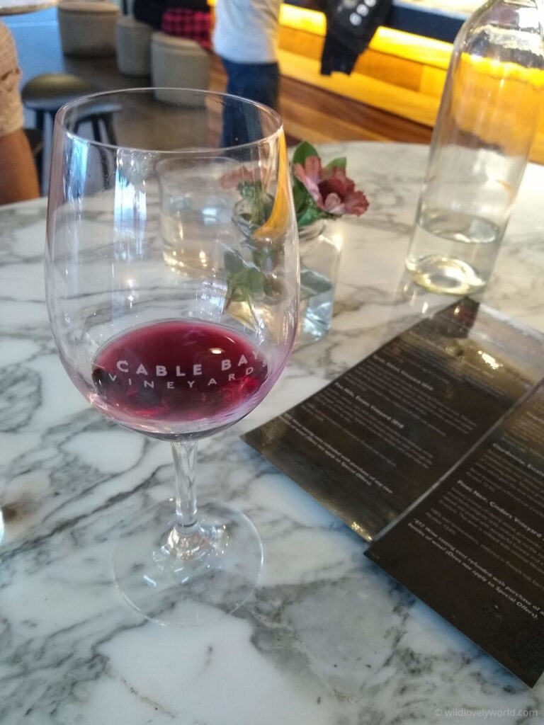 glas of wine on a marble table, the wine has red wine in it and the glass has an etching reading cable bay vineyard. a black menu with white writing is on the table. there is also a small flower in a glass vase with water and a glass bottle of water on the table