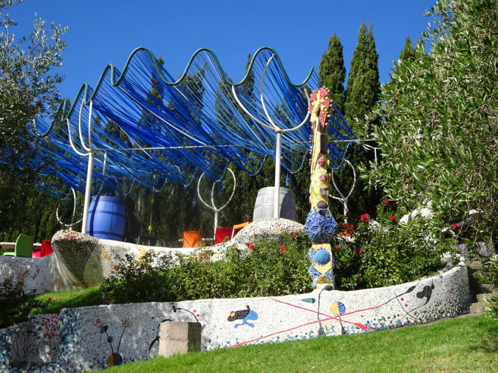 casita miro winery garden terrace, with blue ribbon pergola, and a ceramic mosaic wall in white with spanish artist miro style elements, and a decorative yellow mosaic tower sculpture, surrounded by trees, bushes and flowers