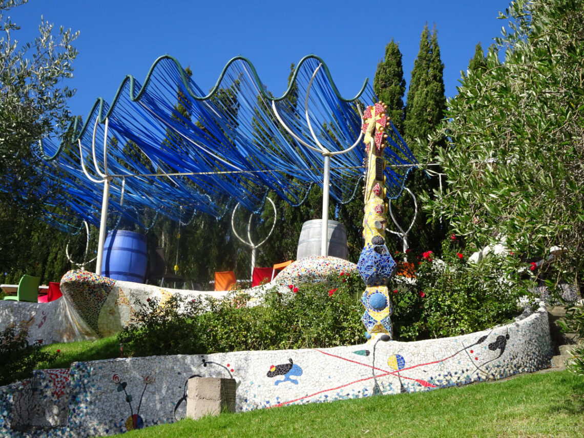 casita miro winery garden terrace, with blue ribbon pergola, and a ceramic mosaic wall in white with spanish artist miro style elements, and a decorative yellow mosaic tower sculpture, surrounded by trees, bushes and flowers