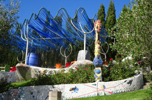 casita miro winery garden terrace, with blue ribbon pergola, and a ceramic mosaic wall in white with spanish artist miro style elements, and a decorative yellow mosaic tower sculpture, surrounded by trees, bushes and flowers