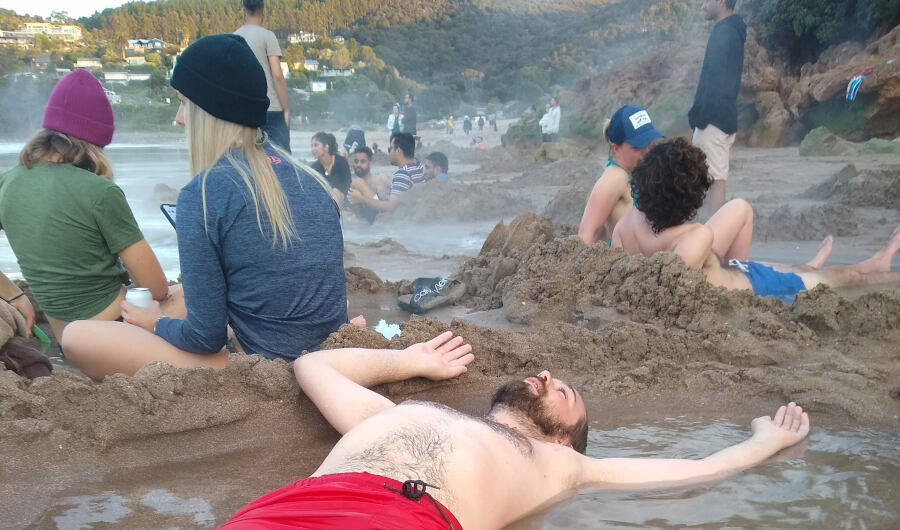 fiachra lying down in the hot water in the sand at coromandel hot water beach, with lots of other people sitting and standing around in the background, with steam rising all around - natural hot springs new zealand