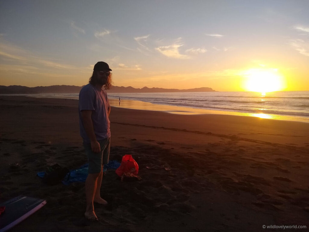 fiachra standing and smiling at the camera, standing on the sandy beach on the left, with the ocean and mountain range in the background and to the right, during sunset at kawhia beach on the west coast of the north island, new zealand