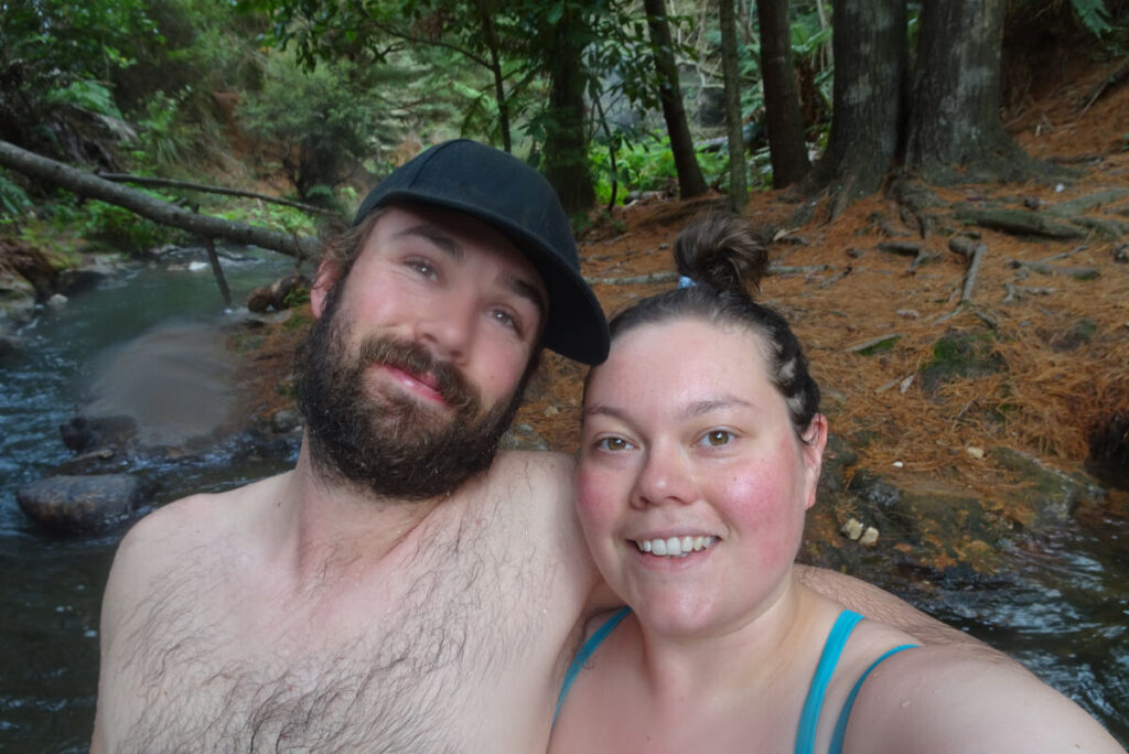 fiachra and lauren smiling at the camera taking a selfie in the hot river at kerosene creek, natural geothermal hot spring in new zealand