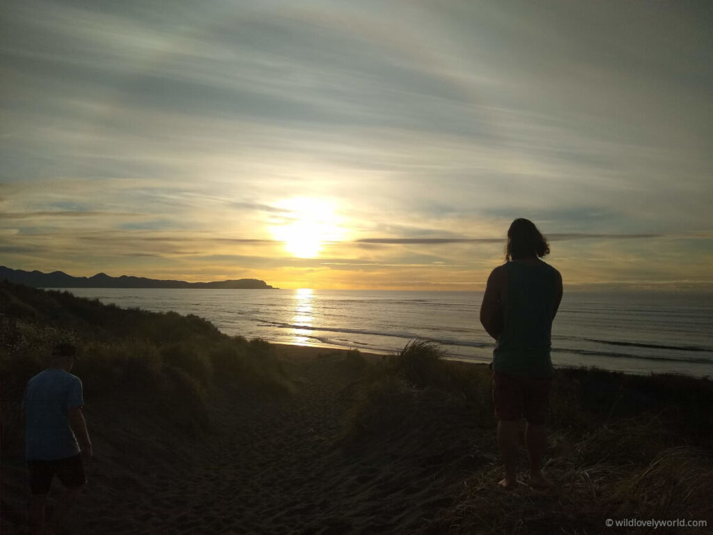 fiachra's silhouette as he looks at the setting sun above the ocean, as he stands on top of a sand dune, above kawhia ocean beach, hot water beach, north island new zealand