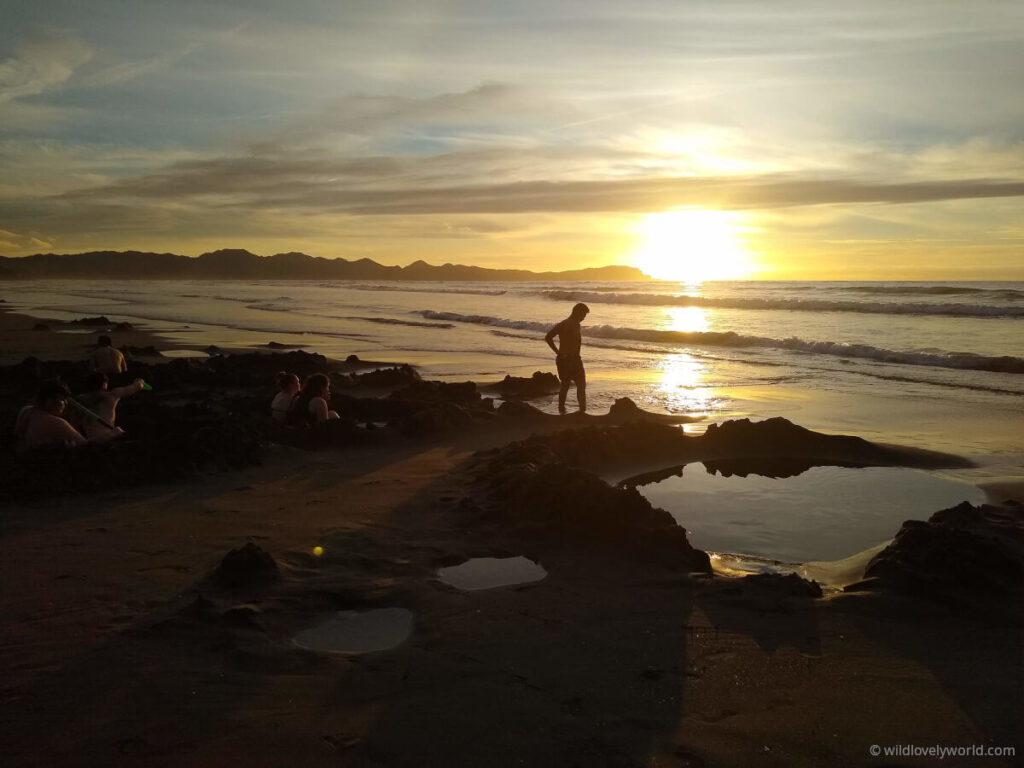 kawhia hot water beach at sunset, with dug holes in the sand filled with water and the sun reflecting on the ocean, and a person standing on the beach and people sitting in the holes.