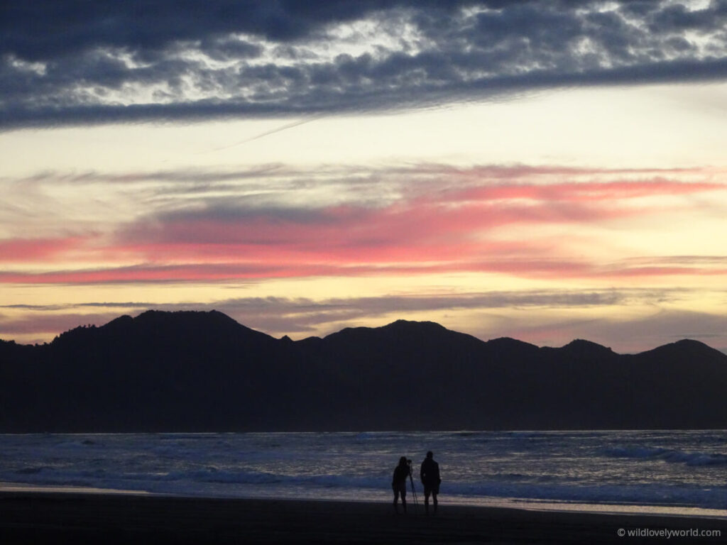 two people standing on the beach, one looking through camera on a tripid, next to the ocean, with black mountains in the distance, and the sky is at sunset and has pink and grey clouds and the sky is a yellow colour, the ocean is dark navy and the sand is black and the people are silhouette