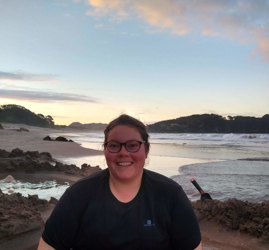 lauren smiling at the camera, wearing glasses and a black t-shirt, sitting in the sand at sunset at Coromandel Hot Water Beach, new zealand