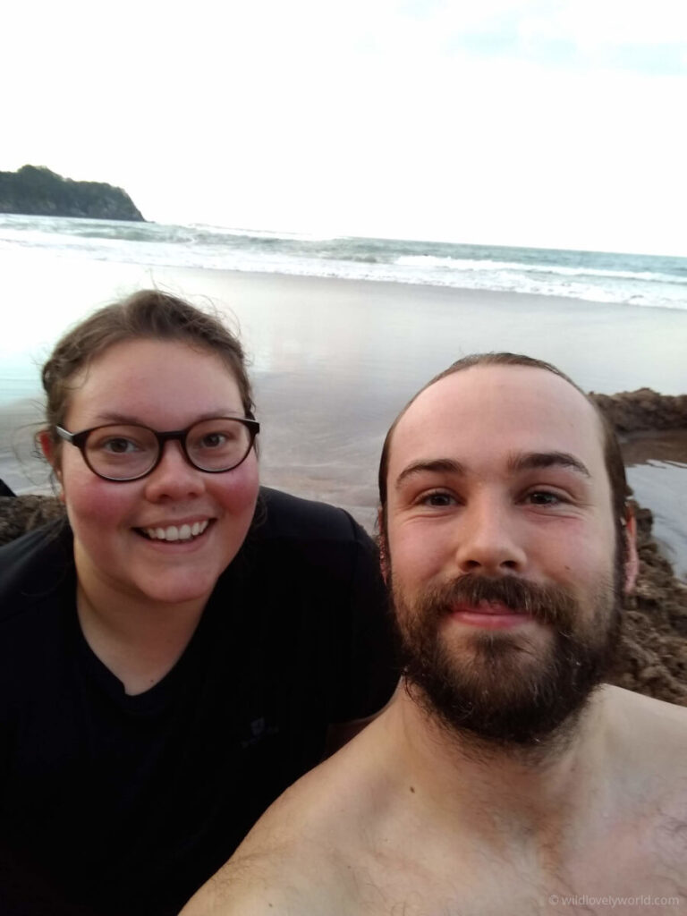 lauren and fiachra smiling at the camera taking a selfie at coromandel hot water beach - they are sitting in a pool dug into the sand, the ocean is behind them