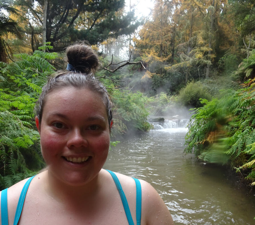 lauren smiling at the camera, wearing swimsuit and hair in bun, standing in the hot river at kerosene creek, surrounded by ferns and trees, with a small waterfall behind her with rising steam - free geothermal hot pools and springs north island new zealand