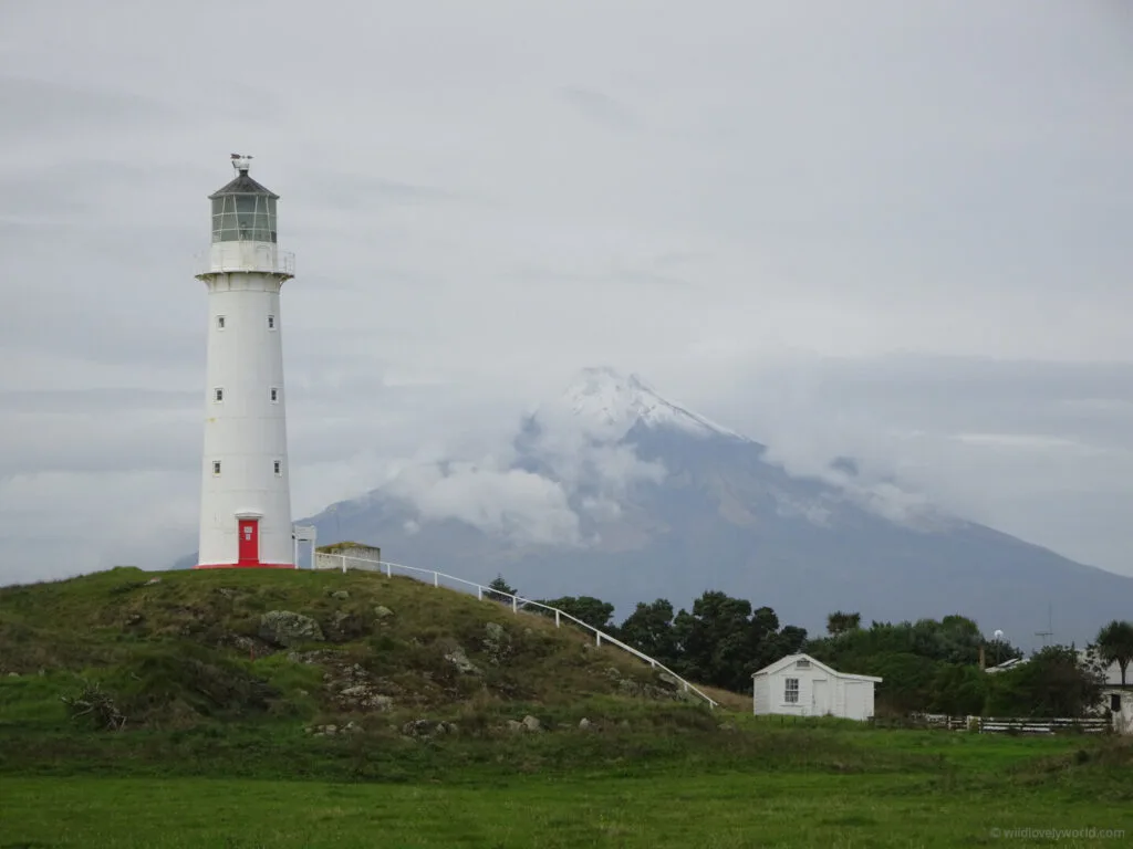 cape egmont lighthouse in the foreground - white with a red door - on a mound with mount taranaki in the background - snow capped pyramid shaped mountain, surrounded by clouds, and a small white farm shed in the middle distance