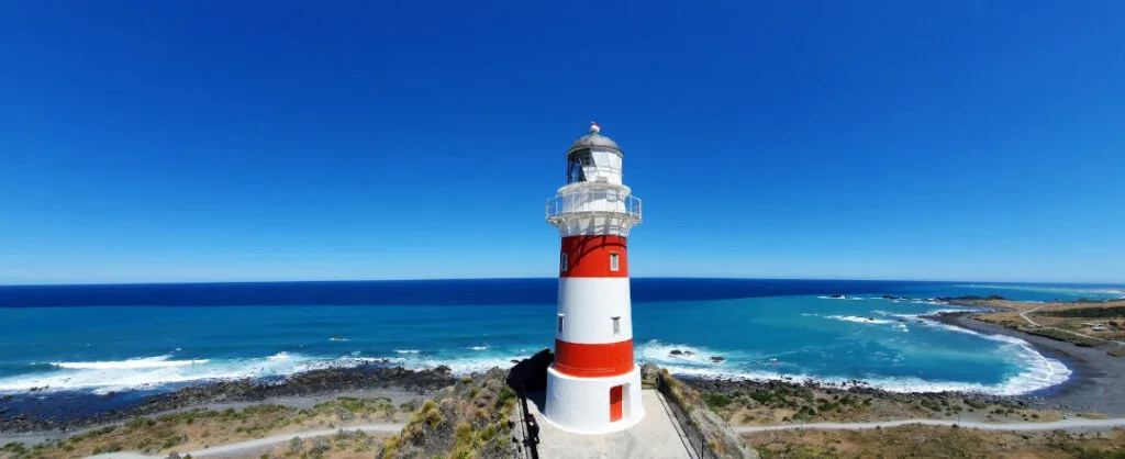 cape palliser lighthouse - red and white striped lighthouse - on a platform - with blue sky and sea with waves and beach and a gravel road along the coastline in new zealand