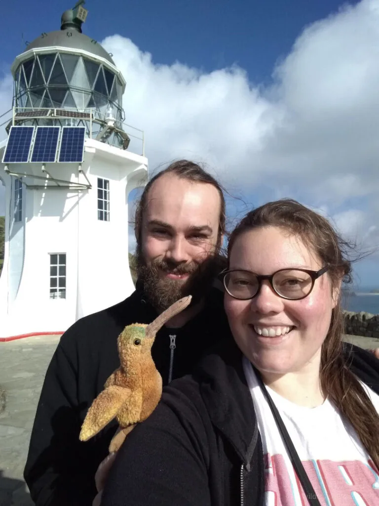 lauren and fiachra smiling and holding up a kiwi beanie baby toy, in front of cape reinga lighthouse in new zealand