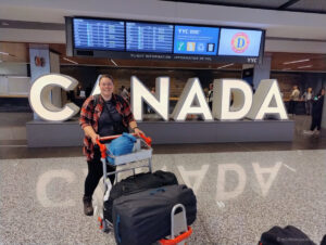 lauren in calgary airport with luggage cart in front of a large canada sign and flight information screens in the background