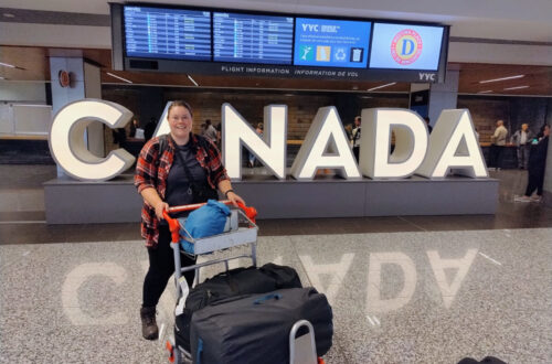 lauren in calgary airport with luggage cart in front of a large canada sign and flight information screens in the background