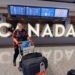 lauren in calgary airport with luggage cart in front of a large canada sign and flight information screens in the background
