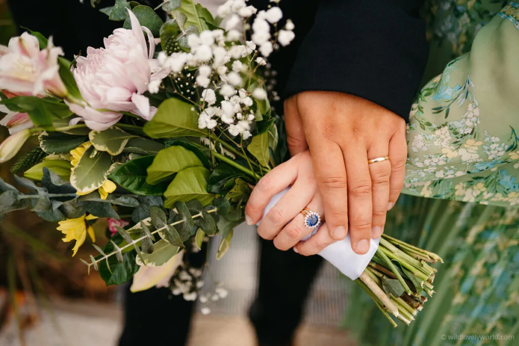 close up of a couples hands with wedding rings, blue engagement ring, holding a flower bouquet