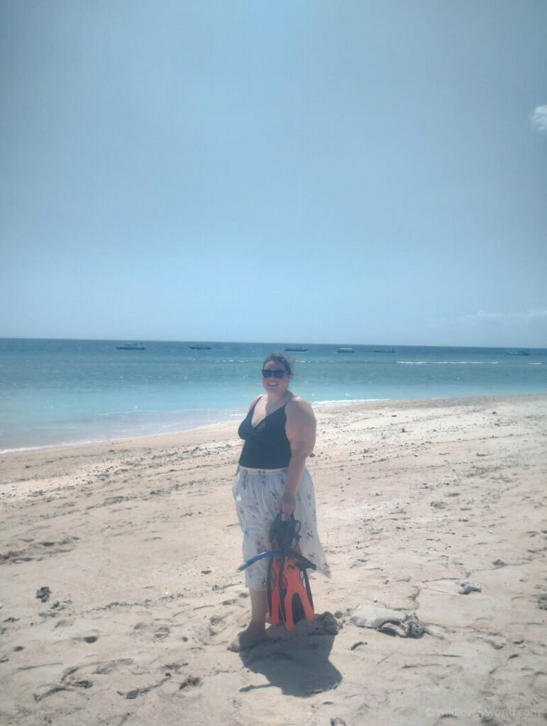 lauren standing on golden gili meno island beach holding snorkelling gear at one of the best snorkelling spots with blue water behind her