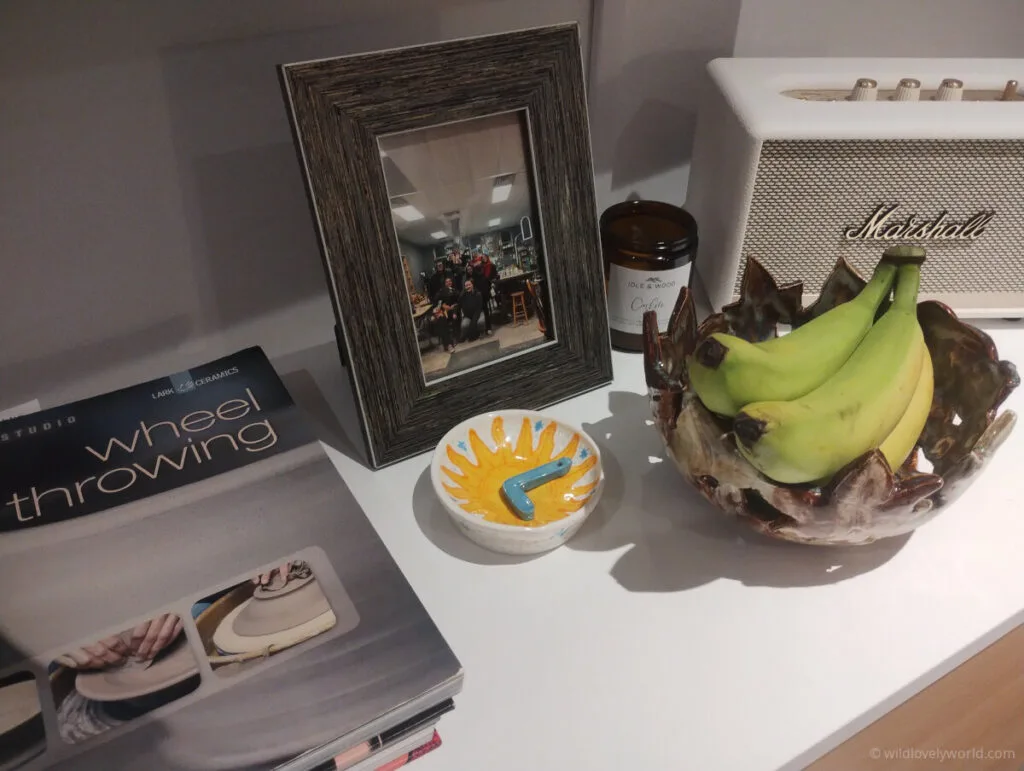 items on a table including a wheel throwing book,a picture frame with photo of friends at a pottery studio, a sun dish with an l in it, a handmade puzzle bowl with bananas
