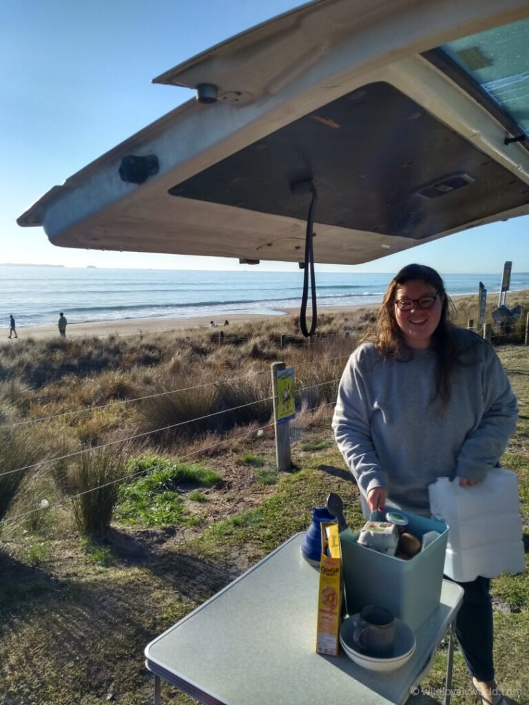 lauren standing at the back of the campervan with breakfast making items with papamoa beach in the background