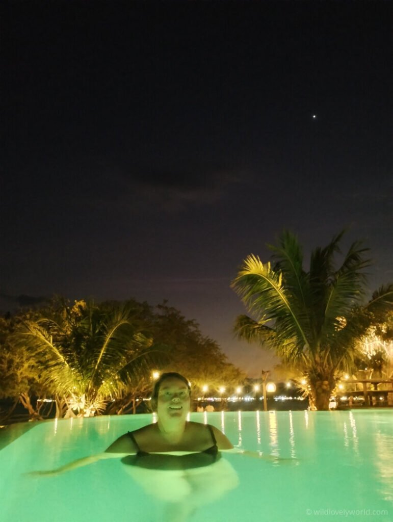 lauren swimming in blue outdoor pool at night with palm trees, fairy lights and star in the sky